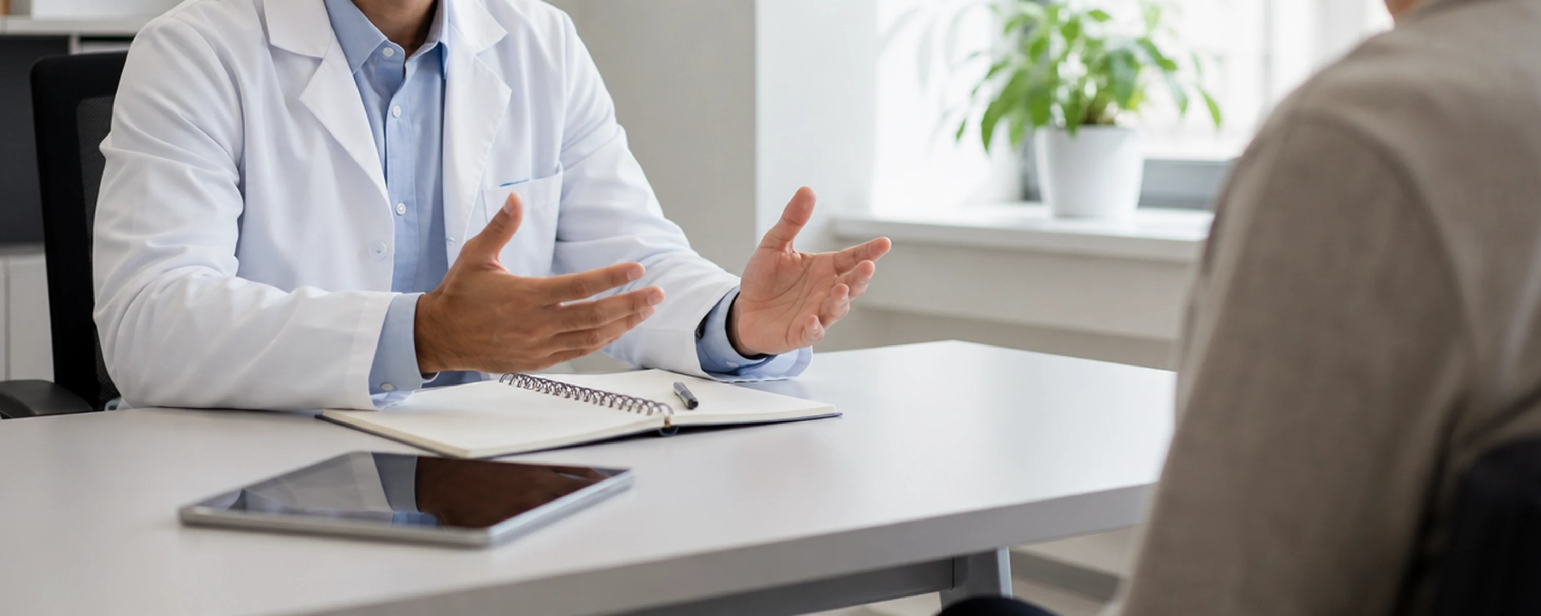Doctor in a white coat gesturing across a clinic desk during a consultation, with a notepad and tablet on the desk and a patient seated opposite
